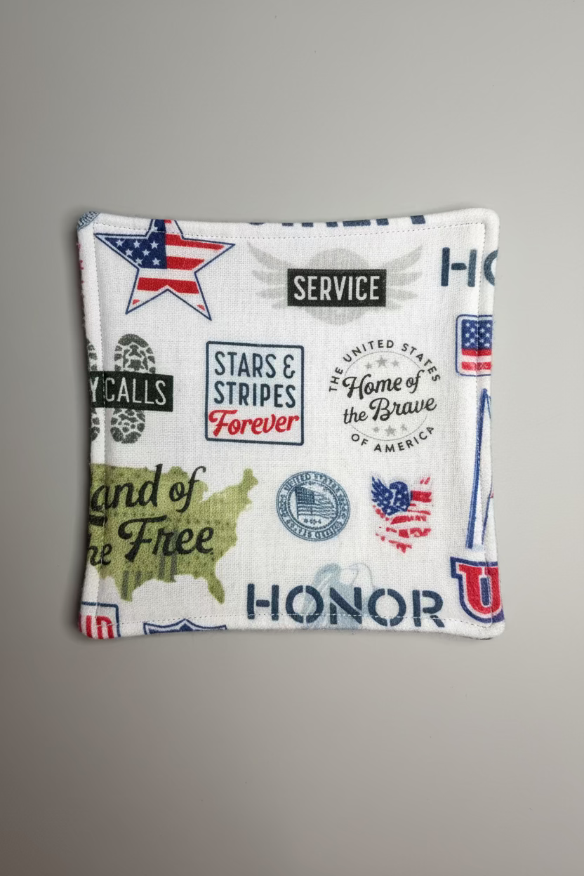 A square white fabric patch with patriotic US-themed graphics, including stars, stripes, the American flag, and text such as 'HONOR,' 'SERVICE,' and 'Home of the Brave,' placed on a gray grid cutting mat.