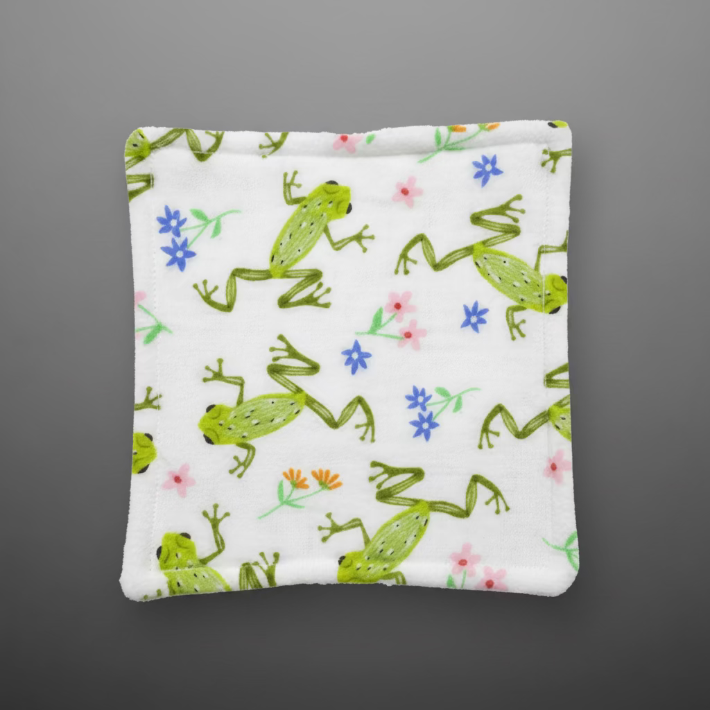 A square white fabric item, likely a coaster or small pad, featuring a green frog and small pink, blue, and orange flower print, placed on a gray grid cutting mat.