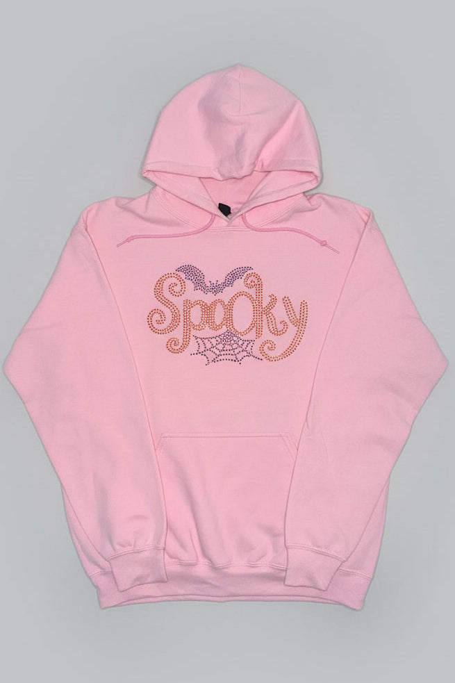 A light pink pullover hoodie with a 'Spooky' rhinestone graphic on the chest, featuring bat wings and a spiderweb design, laid flat on a dark marble surface.