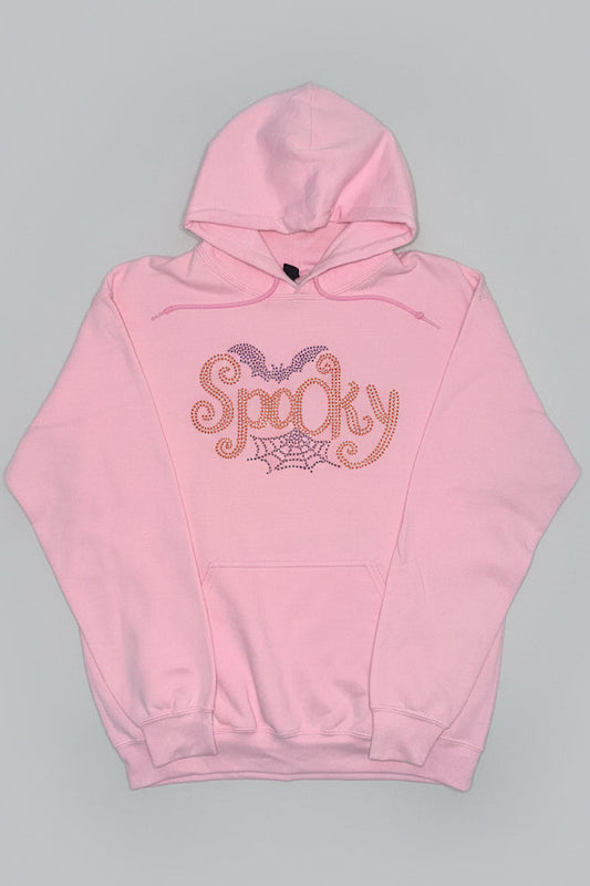 A light pink pullover hoodie with a 'Spooky' rhinestone graphic on the chest, featuring bat wings and a spiderweb design, laid flat on a dark marble surface.
