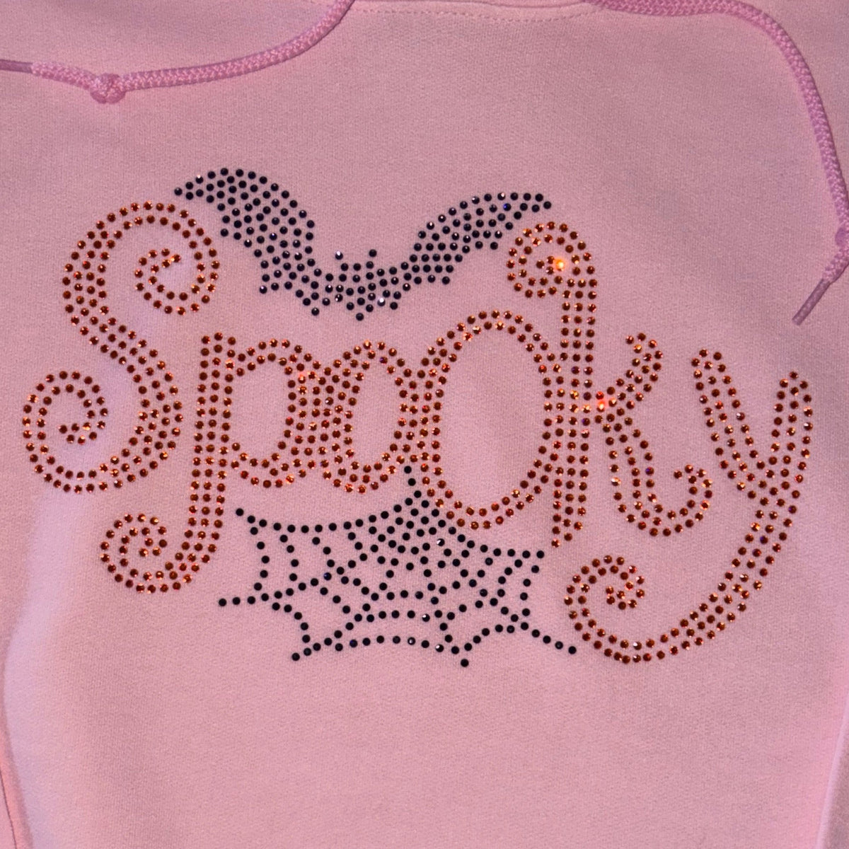 A light pink hoodie featuring a rhinestone-studded 'Spooky' graphic on the chest, complete with a bat and spiderweb design.