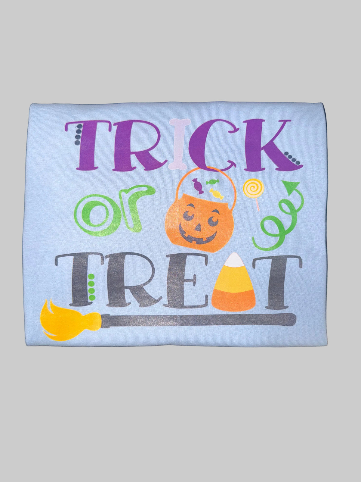 A light blue rectangular fabric item, likely a sweatshirt or t-shirt panel, featuring a colorful Halloween graphic that reads 'TRICK or TREAT' with a jack-o'-lantern, candy corn, broom, and swirls, placed on a dark marbled surface.
