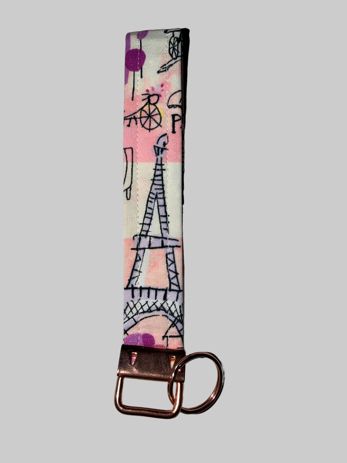 A fabric keychain strap featuring a Paris-themed design with the Eiffel Tower, a bicycle, and a postcard motif on a pink, purple, and cream background, finished with a copper-colored metal clasp and split ring, set against a solid gray background.