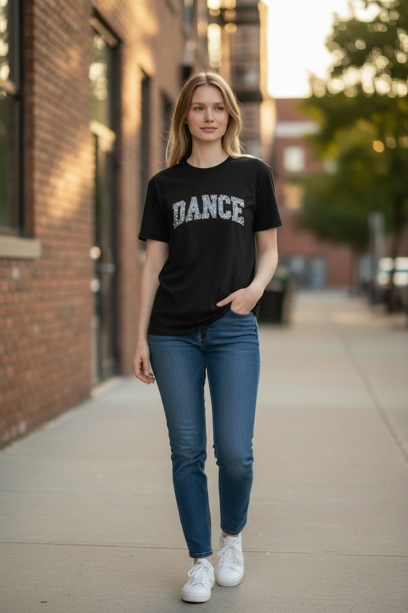 A woman walking on a city sidewalk wearing a black graphic t-shirt with 'DANCE' printed in white across the chest, paired with blue jeans and white sneakers.