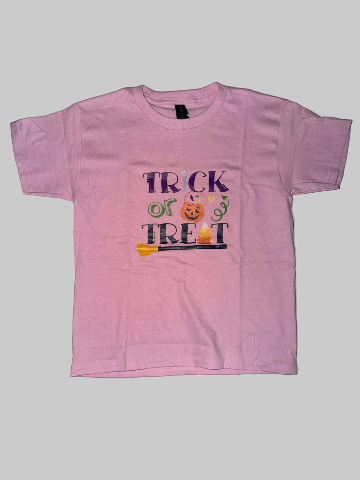 A pink short-sleeve kids' t-shirt with a 'TRICK OR TREAT' graphic, laid flat on a light-colored tiled floor.