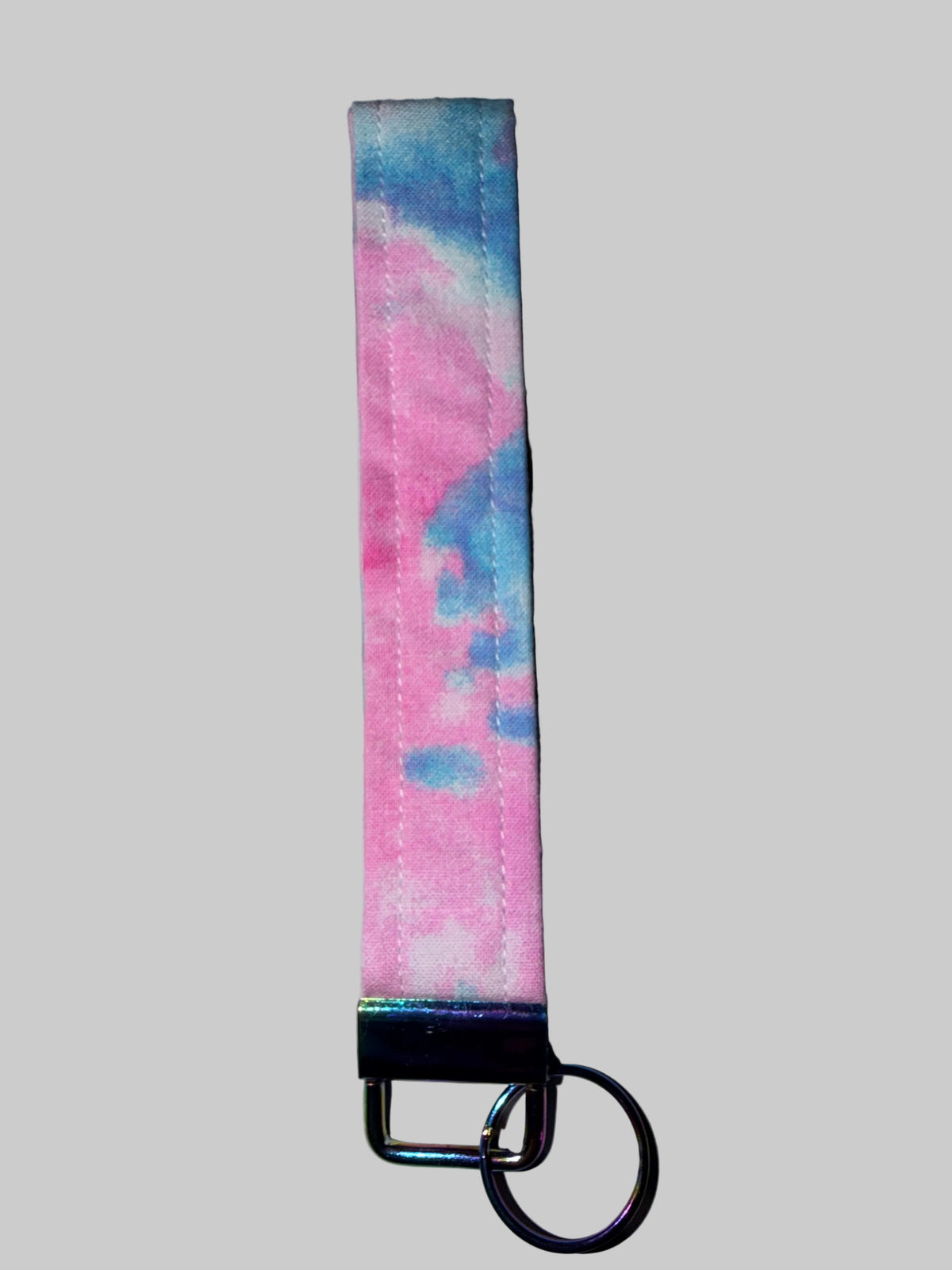 A fabric keychain strap with a pink and blue tie-dye pattern, featuring a black metal clasp and a silver split ring, resting on a white marble-patterned surface.