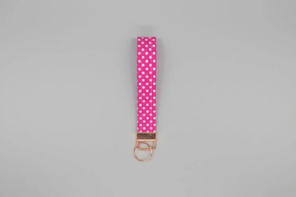 A pink fabric wristlet strap with white polka dots and a rose gold metal key ring clasp, shown against a solid gray background.