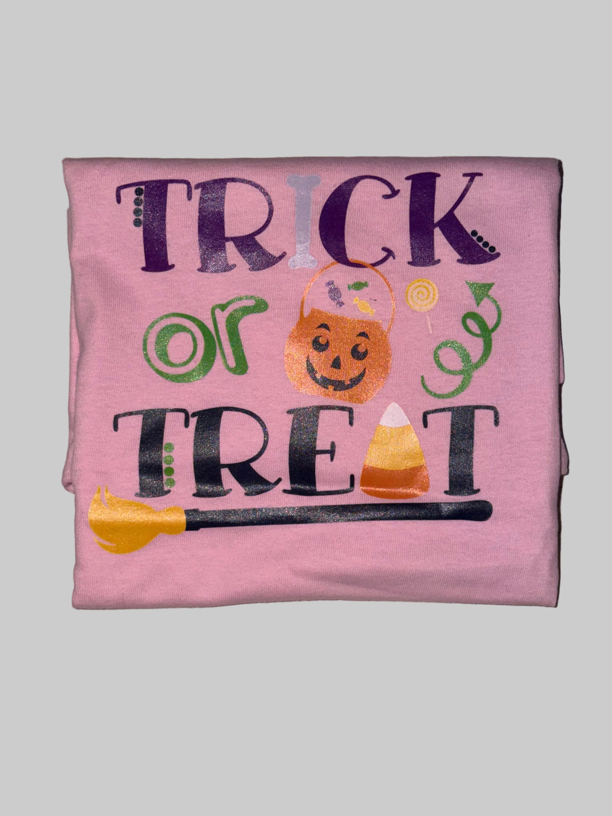 A folded pink t-shirt with a 'Trick or Treat' Halloween graphic, laid flat on a light beige tiled floor.