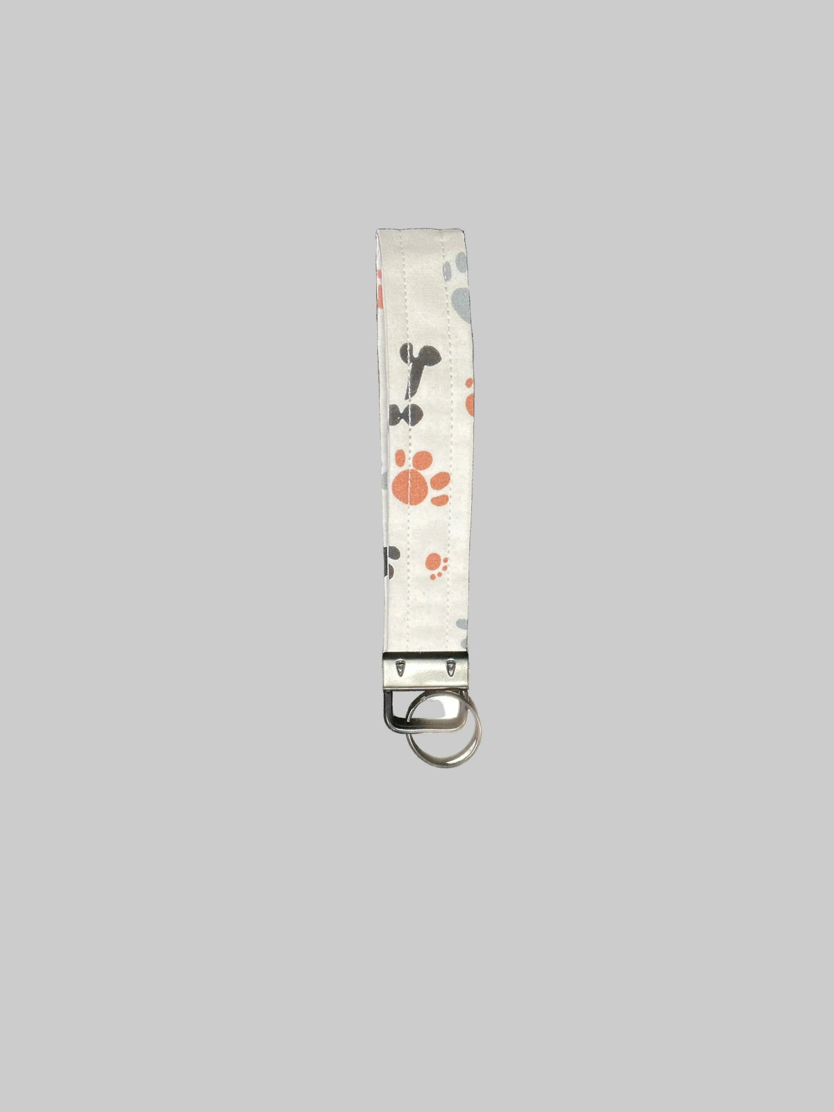 A fabric pet keychain or wristlet strap with a white background, printed with orange paw prints, black and gray bones, and a silver metal clasp with a key ring, shown on a solid gray background.