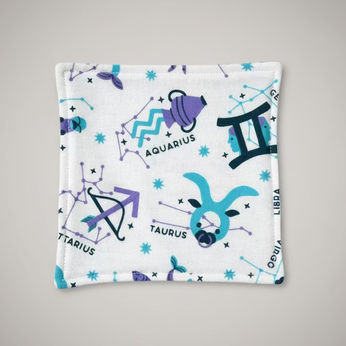 A square white fabric pouch printed with purple and blue zodiac and constellation motifs, including Aquarius, Libra, Taurus, and Scorpio, placed on a gray cutting mat.