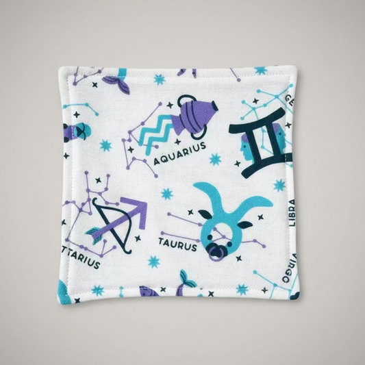 A square white fabric pouch printed with purple and blue zodiac and constellation motifs, including Aquarius, Libra, Taurus, and Scorpio, placed on a gray cutting mat.