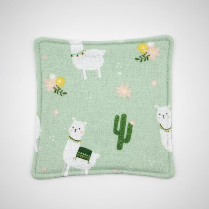 A square light green fabric item, likely a coaster or small mat, featuring embroidered white Llamas, a green cactus, and small yellow and pink flowers, laid flat on a gray grid cutting mat.
